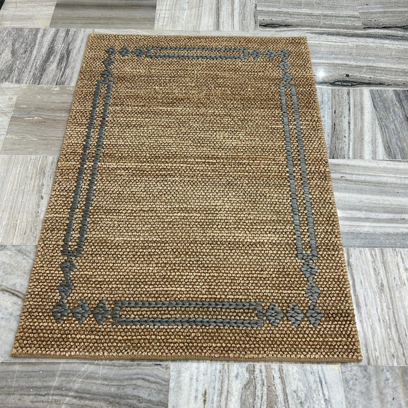 Puede incluir: Alfombra rectangular de yute marr&oacute;n con borde gris. La alfombra tiene una textura tejida y un dise&ntilde;o geom&eacute;trico en los bordes. Adecuada para sal&oacute;n o dormitorio.