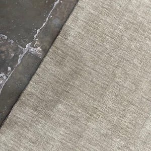 May include: A neutral-toned, textured rug with a subtle pattern. The rug's surface has a woven appearance, and the color is a light, earthy tone. The rug is partially visible, with a contrasting dark, wet surface in the background.