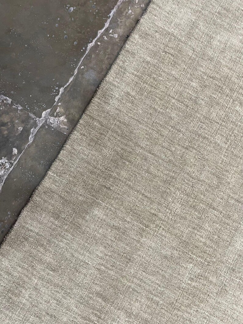 May include: A neutral-toned, textured rug with a subtle pattern. The rug's surface has a woven appearance, and the color is a light, earthy tone. The rug is partially visible, with a contrasting dark, wet surface in the background.