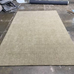 May include: A large, rectangular area rug in a neutral beige color. The rug has a textured surface and is placed on a concrete floor. Other rugs and materials are visible in the background.