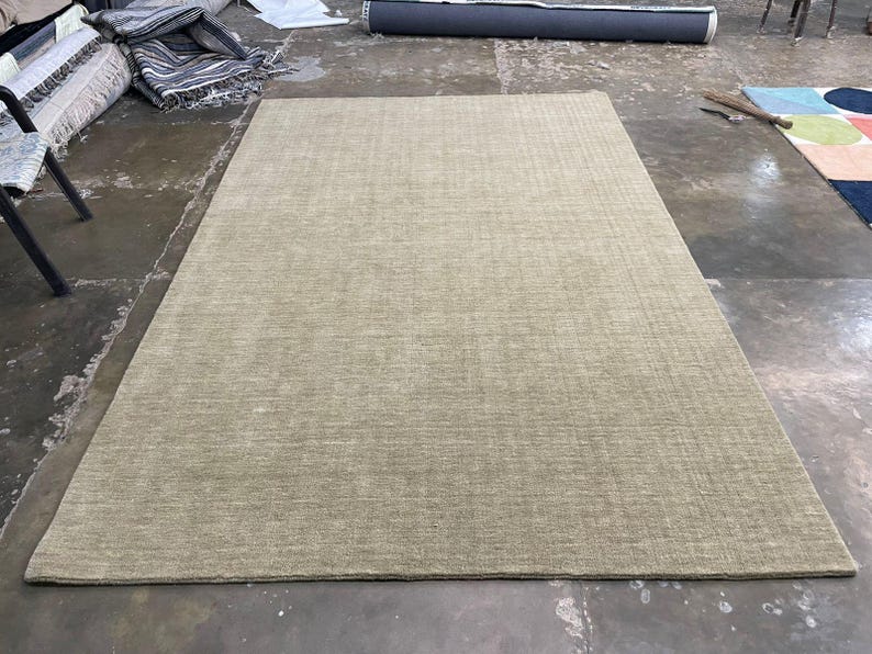May include: A large, rectangular area rug in a neutral beige color. The rug has a textured surface and is placed on a concrete floor. Other rugs and materials are visible in the background.