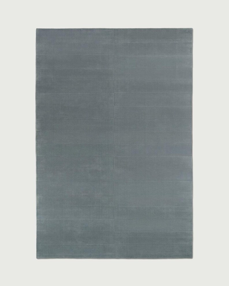 Puede incluir: Una alfombra rectangular de color azul pizarra s&oacute;lido. La superficie presenta una serie de l&iacute;neas horizontales paralelas y hundidas que crean un sutil patr&oacute;n de cuadr&iacute;cula en toda la longitud y anchura del revestimiento textil.