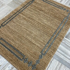 Puede incluir: Una alfombra rectangular tejida en color beige natural con un borde gris. El borde presenta l&iacute;neas paralelas y elementos decorativos en las esquinas. La alfombra tiene una superficie texturizada.