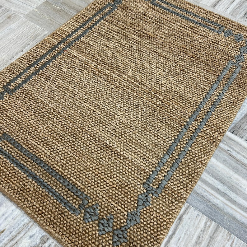 Puede incluir: Una alfombra rectangular tejida en color beige natural con un borde gris. El borde presenta l&iacute;neas paralelas y elementos decorativos en las esquinas. La alfombra tiene una superficie texturizada.