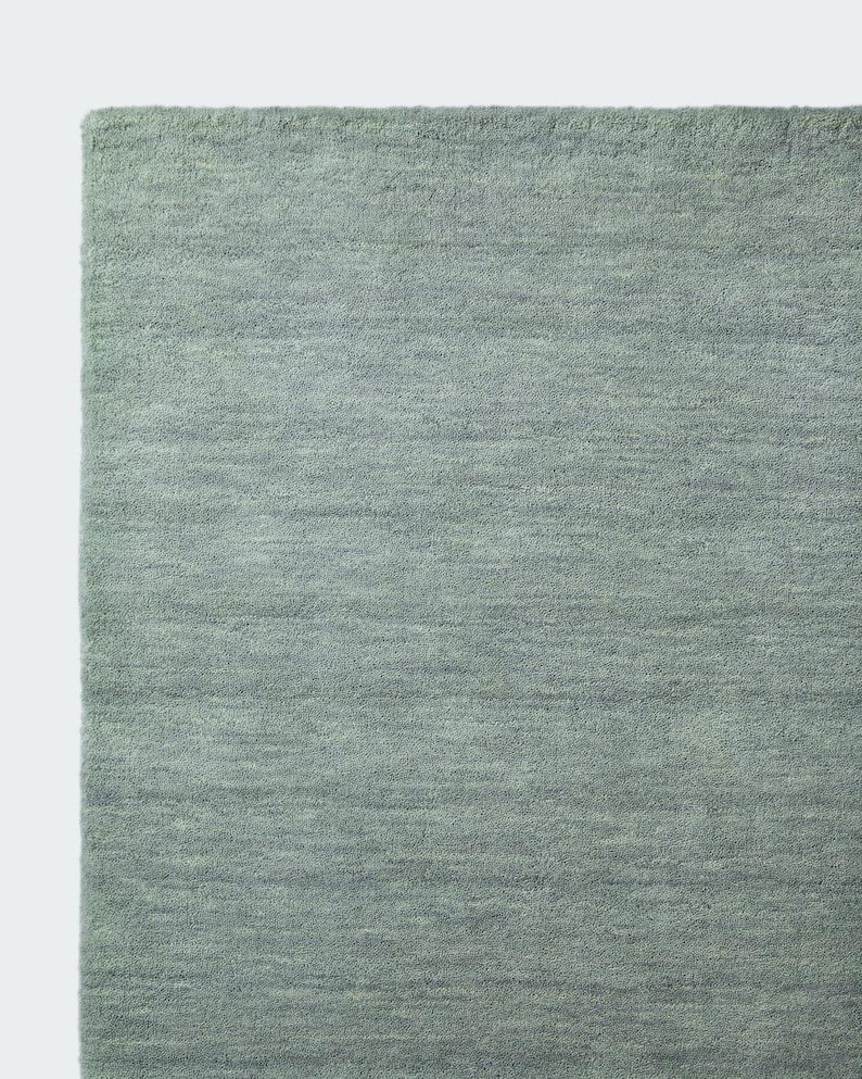 May include: A rectangular area rug in a solid sage green colour. The rug features a dense, short-pile texture with subtle horizontal variations in the weave, creating a soft, uniform appearance across the entire surface of the floor covering.