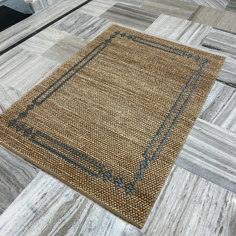 Puede incluir: Alfombra rectangular tejida en tonos beige y marr&oacute;n. Presenta un borde gris oscuro con elementos decorativos. La alfombra tiene una superficie texturizada y est&aacute; colocada sobre un suelo efecto m&aacute;rmol.