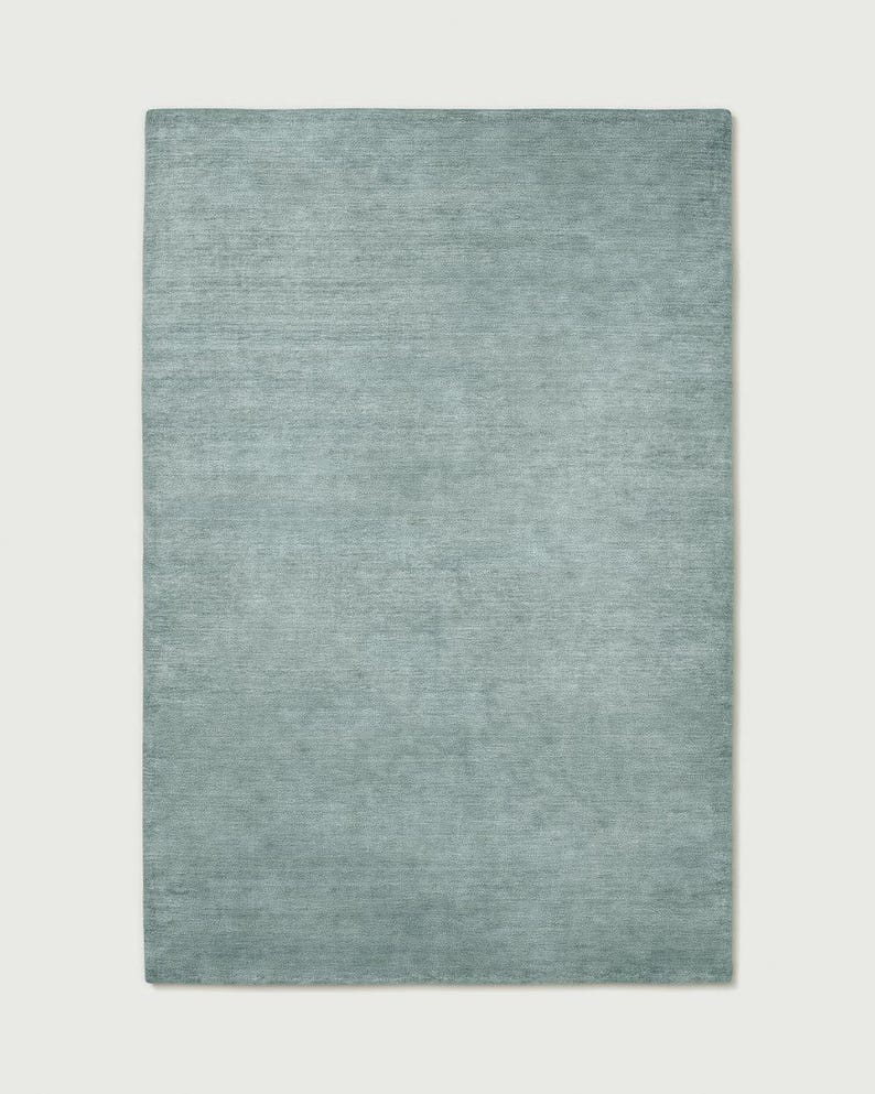 May include: A rectangular area rug in a solid, muted sage green colour. The surface features a subtle, horizontal linear texture that runs across the entire length of the rug, creating a soft, uniform appearance with slight variations in tone.