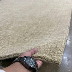 May include: A large, rectangular beige rug with a textured surface. The rug is held at the corner, showcasing its thickness and color. The rug appears to be made of a soft material.