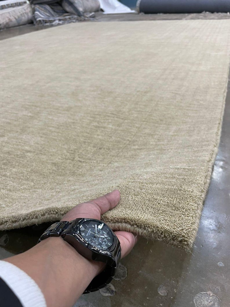 May include: A large, rectangular beige rug with a textured surface. The rug is held at the corner, showcasing its thickness and color. The rug appears to be made of a soft material.