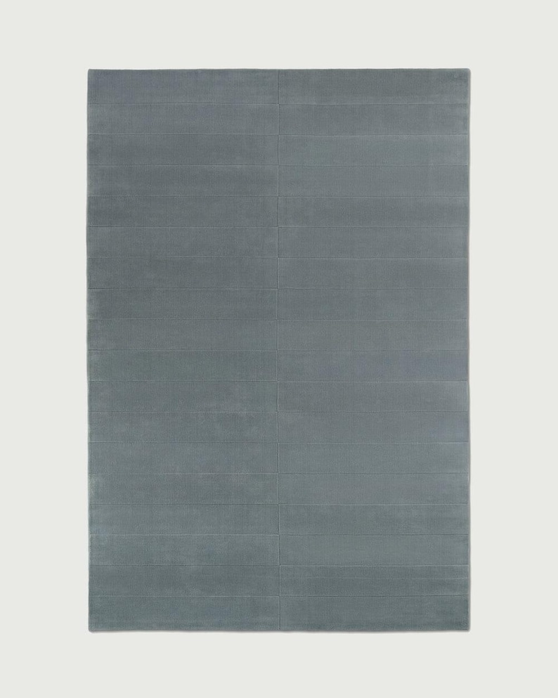 Puede incluir: Una alfombra rectangular de color azul pizarra s&oacute;lido. La superficie presenta una serie de l&iacute;neas horizontales paralelas y hundidas que crean un sutil patr&oacute;n de cuadr&iacute;cula en toda la longitud y anchura del revestimiento textil.