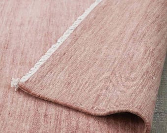 May include: A rolled-up, dusty rose-colored rug with a textured weave. The rug's edge reveals a white fringe. The rug is made of a woven material and is displayed on a wooden surface.