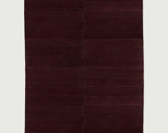 Puede incluir: Una alfombra rectangular de color burdeos oscuro y profundo. La alfombra presenta una serie de líneas horizontales paralelas que crean una apariencia segmentada y panelada en toda la superficie, con una costura vertical en el centro.