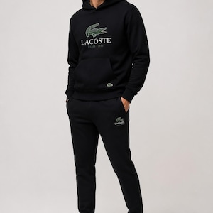 May include: A person wearing a black Lacoste hoodie and matching sweatpants. The hoodie features the Lacoste logo and "Paris 1933" text. White sneakers complete the outfit. The Lacoste logo is also on the sweatpants.