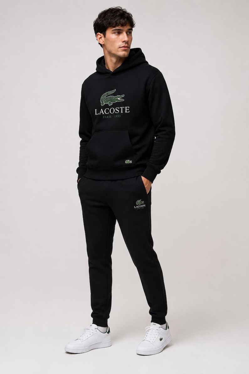 May include: A person wearing a black Lacoste hoodie and matching sweatpants. The hoodie features the Lacoste logo and "Paris 1933" text. White sneakers complete the outfit. The Lacoste logo is also on the sweatpants.