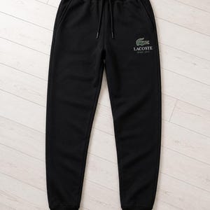May include: Black sweatpants with an elastic waistband and drawstring. The pants feature a small green and white Lacoste logo on the upper left leg. The logo includes the text "Lacoste Paris 1933".