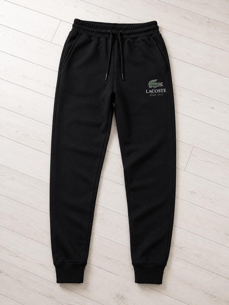 May include: Black sweatpants with an elastic waistband and drawstring. The pants feature a small green and white Lacoste logo on the upper left leg. The logo includes the text "Lacoste Paris 1933".