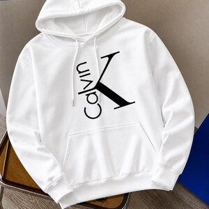 May include: White hooded sweatshirt with a front pocket and drawstring. The sweatshirt features the black text "Calvin K" on the front. The hoodie is made of a soft material.