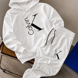 May include: White hoodie and sweatpants set. The hoodie features a drawstring hood and a front pocket. The set has the word "Calvin" and a "K" logo in black. The sweatpants have an elastic waistband and drawstring.