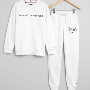 Puede incluir: Conjunto de sudadera y pantalón de chándal blancos. La sudadera tiene el texto "TOMMY HILFIGER" en azul marino. El pantalón de chándal tiene cintura con cordón y el mismo texto en azul marino. Ambos artículos tienen el logotipo de Tommy Hilfiger.