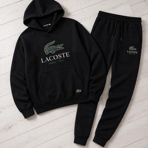 May include: Black Lacoste hoodie and matching sweatpants set. The hoodie features a green crocodile logo and the text "Lacoste Paris 1933". The sweatpants also have the Lacoste logo. The set is displayed on a light wood grain surface.