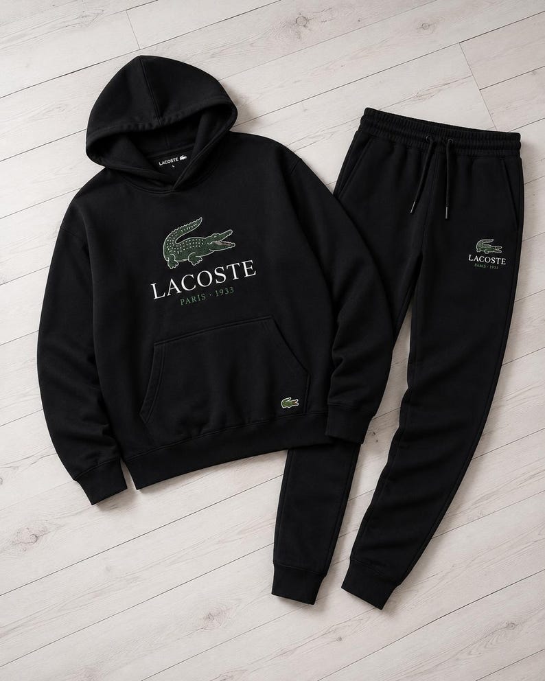 May include: Black Lacoste hoodie and matching sweatpants set. The hoodie features a green crocodile logo and the text "Lacoste Paris 1933". The sweatpants also have the Lacoste logo. The set is displayed on a light wood grain surface.
