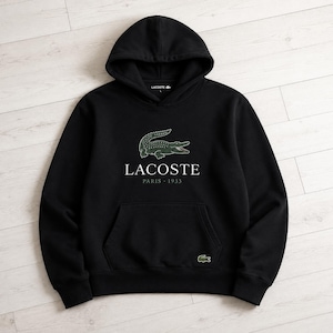 May include: Black hooded sweatshirt with a kangaroo pocket and the Lacoste logo. The logo features a green crocodile above the text "LACOSTE PARIS 1933". A small green crocodile logo is also on the lower right side of the sweatshirt.