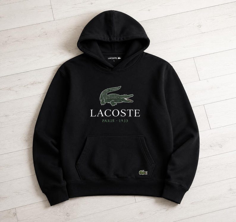 May include: Black hooded sweatshirt with a kangaroo pocket and the Lacoste logo. The logo features a green crocodile above the text "LACOSTE PARIS 1933". A small green crocodile logo is also on the lower right side of the sweatshirt.