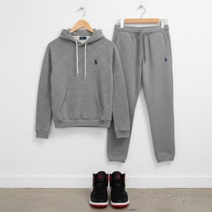 May include: A gray hoodie and matching sweatpants set with a small navy blue logo. The hoodie has a front pocket and white drawstrings. Black and red sneakers with white soles are at the bottom.