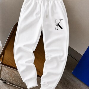 May include: White sweatpants with a drawstring waist and ribbed cuffs. The pants feature the black text "Calvin" and a "K" logo on the left leg. The pants are made of a soft, comfortable material.