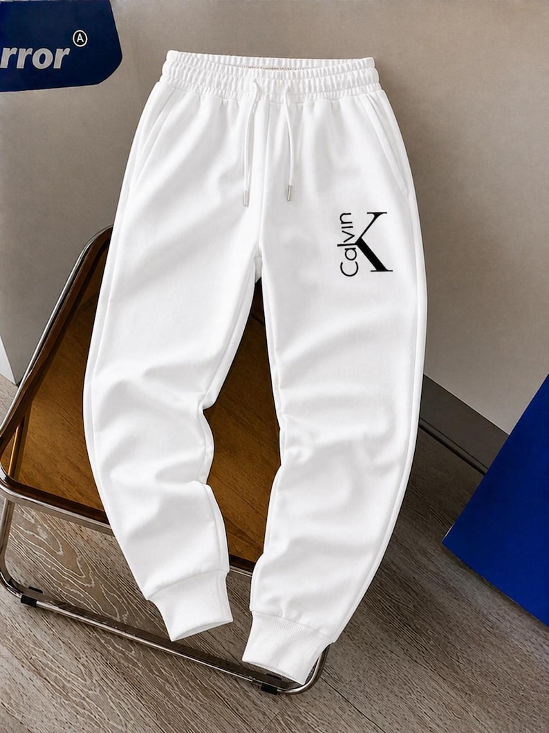 May include: White sweatpants with a drawstring waist and ribbed cuffs. The pants feature the black text "Calvin" and a "K" logo on the left leg. The pants are made of a soft, comfortable material.