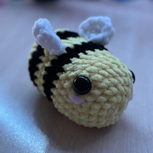 Op de afbeelding: Een handgemaakte gehaakte bij met een geel en zwart gestreept lichaam, witte vleugels en grote zwarte ogen. De bij is een zacht, pluche speelgoed, perfect voor kinderen of als decoratie.