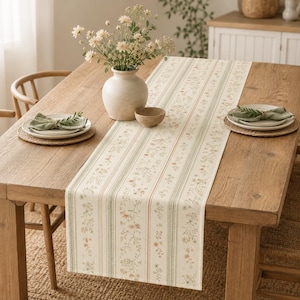 May include: A long rectangular table runner featuring a cream-colored fabric with vertical stripes and delicate floral patterns in muted green, brown, and orange tones, centered on a wooden dining table with place settings and a flower vase.