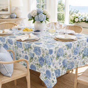May include: A white tablecloth featuring a repeating pattern of blue hydrangea flowers, green leaves, starfish, and seashells. The fabric is draped over a dining table set with white plates, napkins, glassware, and a central vase of hydrangeas.