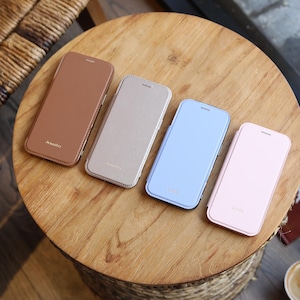 Könnte beinhalten: Vier Leder-Handyhüllen in Braun, Beige, Hellblau und Rosa, jeweils mit personalisiertem Namen in goldfarbenen Buchstaben. Die Hüllen sind auf einem Holztisch angeordnet und zeigen eine Vielzahl von Farben und Stilen.