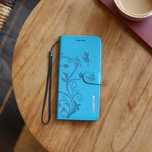 Könnte beinhalten: Türkisfarbene Handyhülle mit Schmetterlings- und Blumenmuster. Die Hülle hat eine Handschlaufe und einen Magnetverschluss. Das Design ist auf der Vorderseite geprägt. Der Text "LSZSHOP" ist unten rechts aufgedruckt.