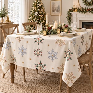 Op de afbeelding: Een crèmekleurig rechthoekig tafelkleed met een herhalend patroon van ingewikkelde sneeuwvlokken, hulstbladeren en kleine rode bessen. De stof ligt over een eettafel, met gouden steraccenten verspreid tussen de grotere motieven.