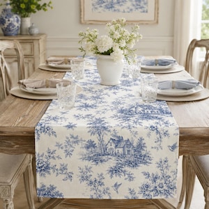 May include: A white table runner featuring a blue toile de jouy pattern with illustrations of houses, trees, and birds. The runner is placed on a wooden dining table set with white plates, light blue napkins, and clear glasses around a white vase.
