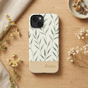 Può includere: Custodia per telefono con sfondo color crema e motivo a foglie verdi. La sezione inferiore è in legno chiaro con il nome "Jessica" inciso. La custodia è circondata da fiori e un panno beige.