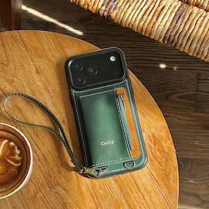 May include: Green leather phone case with a brown accent and a zipper pocket. The case has a wrist strap and the name "Cathy" is printed on the front. The phone case is on a wooden table.