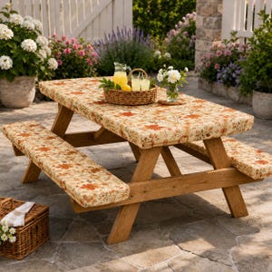May include: A wooden picnic table with attached benches, featuring thick, cushioned pads with a floral pattern in shades of orange, brown, and cream. A woven basket holds a glass pitcher of lemonade and two glasses on the tabletop.