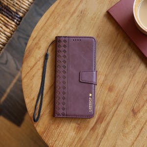 Könnte beinhalten: Eine weinrote Leder-Handyhülle mit einem Webmuster auf der linken Seite und einem Magnetverschluss. Die Hülle hat eine Handschlaufe und den Text "LSZSHOP" mit einem Herzsymbol. Die Hülle liegt auf einem Holztisch.