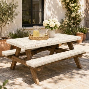 May include: A wooden picnic table featuring a cream-colored surface and bench covers decorated with a delicate floral pattern. The table includes a woven tray holding a glass pitcher of lemonade and a white vase filled with white hydrangea flowers.