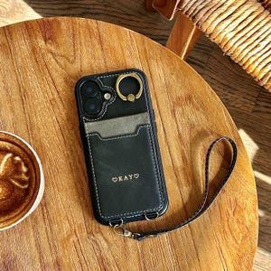 May include: Black phone case with a cardholder and a gold ring grip. The case has a strap and the word "Okay" with a heart symbol is printed on the front. The case is on a wooden table next to a cup of coffee.