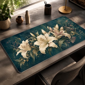 May include: A rectangular desk mat featuring a deep teal background with a central illustration of white lilies and delicate foliage. The floral design spans the length of the mat, showcasing blooming lily petals and green leaves in a soft, painterly style.