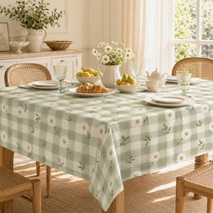 Può includere: Un tavolo da pranzo quadrato coperto da una tovaglia a quadri verde chiaro e bianca con un motivo a margherite bianche. Il tavolo è apparecchiato con piatti bianchi, bicchieri trasparenti, una brocca in ceramica e frutta fresca.