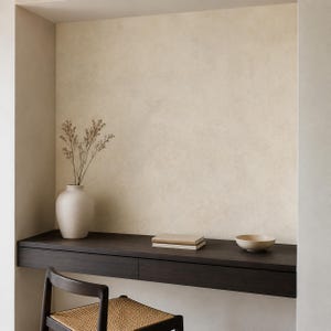 Könnte beinhalten: Ein dunkelbrauner Schwebeschreibtisch mit zwei Schubladen, darauf eine matte cremeweiße Vase mit getrockneten Zweigen, ein Stapel aus zwei beigen Büchern und eine kleine passende Schale. Ein dunkelbrauner Stuhl mit geflochtener Sitzfläche steht davor.