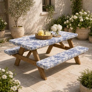 Puede incluir: Una mesa de picnic de madera con bancos integrados que tienen cojines con estampado floral azul y blanco. Sobre la mesa hay una bandeja redonda de mimbre con una jarra de cristal de limonada y dos vasos, junto a un jarrón con flores blancas.