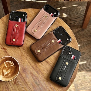 May include: Four leather phone cases in red, pink, brown, and black, each with a card slot and a strap. The cases have gold-coloured button accents and a striped fabric detail. The red and black cases have the name "Lyuda" printed on them, and the brown case has "J. Simon".