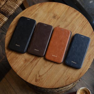 May include: Four leather phone cases in black, brown, tan, and navy blue are arranged on a wooden table. Each case has a name printed on it in gold lettering. The cases are closed and appear to be for a smartphone.
