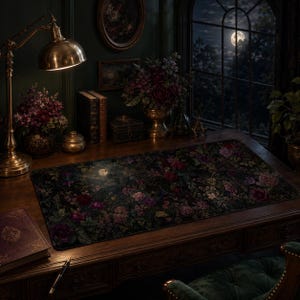 May include: A rectangular desk mat featuring a dark, moody floral pattern with deep red, purple, and green botanical elements. The mat lies flat on a wooden desk surface, showcasing a dense arrangement of blooming flowers and foliage across its surface.
